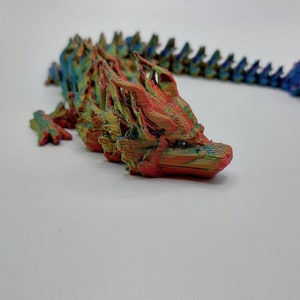May include: A multi-colored 3D printed dragon figurine with a long, segmented tail. The dragon is mostly red and orange with hints of green and blue.