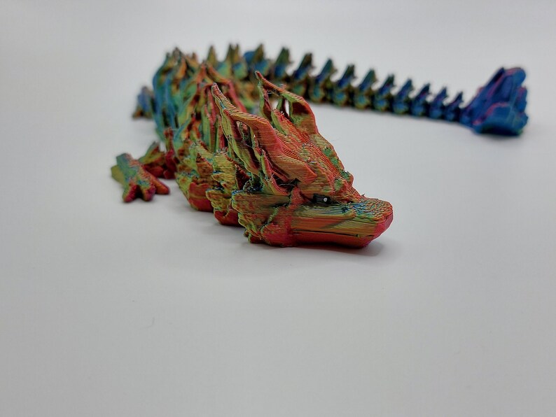 May include: A multi-colored 3D printed dragon figurine with a long, segmented tail. The dragon is mostly red and orange with hints of green and blue.