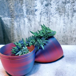 May include: A green and blue 3D printed dragon figurine inside a red and blue ceramic pot. The pot is split in half, with the dragon figurine resting inside one half.