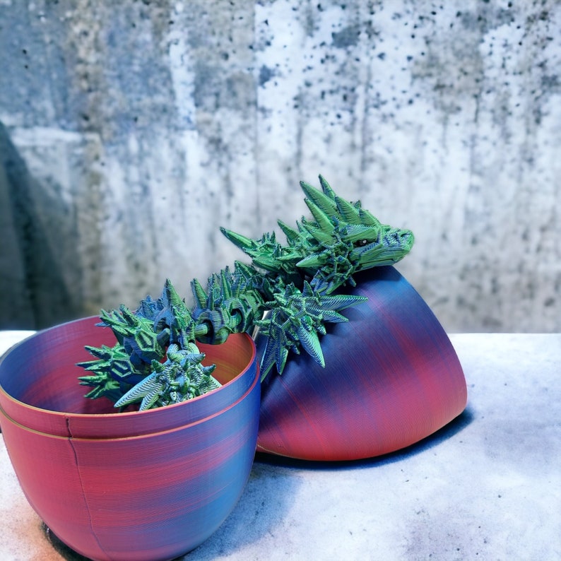 May include: A green and blue 3D printed dragon figurine inside a red and blue ceramic pot. The pot is split in half, with the dragon figurine resting inside one half.