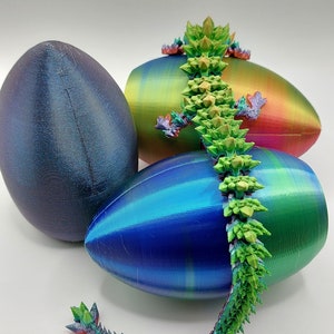 May include: A 3D printed, iridescent green and red dragon figurine with spiky scales, perched on top of three iridescent Easter eggs. The eggs are blue, green, and purple.