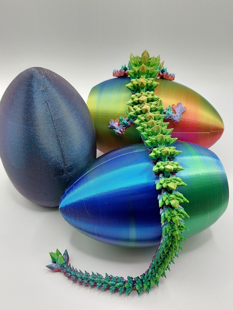 May include: A 3D printed, iridescent green and red dragon figurine with spiky scales, perched on top of three iridescent Easter eggs. The eggs are blue, green, and purple.