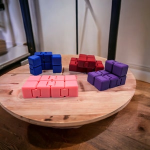 May include: Four colorful fidget toys made of plastic. The toys are in the shape of cubes and are arranged on a wooden surface. The toys are blue, red, purple, and pink.