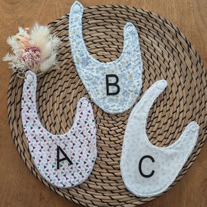 Peut inclure: Trois bavoirs pour bébé avec fermetures à pression. Le bavoir A présente un motif de triangles roses et gris. Le bavoir B a un imprimé floral bleu et jaune. Le bavoir C est blanc avec un petit motif floral rose. Chaque bavoir porte une lettre noire A, B ou C.