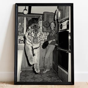 Puede incluir: Impresión enmarcada en blanco y negro que representa una escena de cocina surrealista. Un tigre parece estar entrando en la cocina, mientras una persona está cerca sosteniendo una sartén. La imagen tiene una estética vintage.