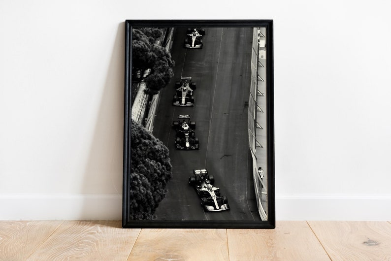 May include: Black and white photograph of five race cars driving on a winding road. The cars are in a line, with the lead car at the top of the image and the last car at the bottom. The road is surrounded by trees and a barrier.