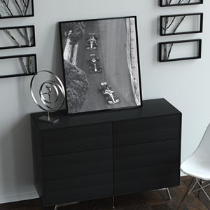 May include: A black and white photograph of race cars on a track, framed in a black frame and hanging on a white wall above a black dresser with six drawers. A silver metal sculpture sits on the dresser.