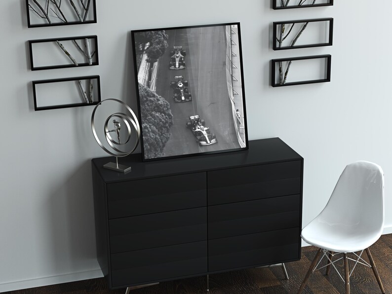 May include: A black and white photograph of race cars on a track, framed in a black frame and hanging on a white wall above a black dresser with six drawers. A silver metal sculpture sits on the dresser.