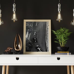 May include: A white desk with two drawers and wooden legs. The desk has a black and white photo of race cars on a track in a gold frame. There are four hanging light bulbs with clear glass shades and a small bronze sculpture on the desk.