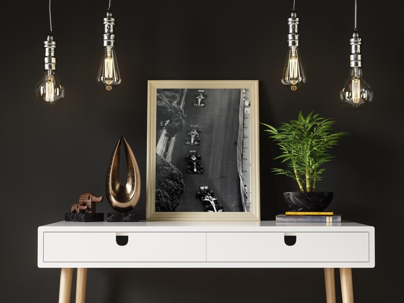 May include: A white desk with two drawers and wooden legs. The desk has a black and white photo of race cars on a track in a gold frame. There are four hanging light bulbs with clear glass shades and a small bronze sculpture on the desk.