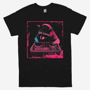 Puede incluir: Camiseta negra con un diseño gráfico en rosa y azul neón de un mapache con auriculares mezclando música en una mesa de mezclas. El diseño está dentro de un cuadrado rosa.