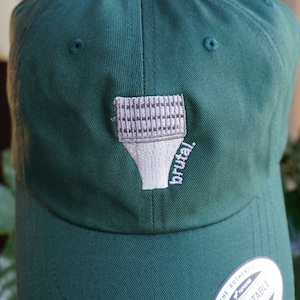 Könnte beinhalten: Eine dunkelgrüne Baseballkappe mit einem gestickten weißen Pinsel und dem Wort "brutal" auf der Vorderseite. Die Kappe hat einen verstellbaren Riemen auf der Rückseite mit einem weißen Etikett, auf dem "The Authentic Yupoong Adjustable" steht.