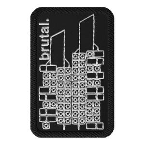 May include: Black rectangular patch with a white embroidered design of a building and the word "brutal." The patch has a black border. The building design features a series of rectangular shapes and three tall, thin structures.