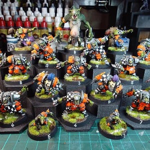 May include: A collection of hand-painted miniature orc figurines in orange and black checkered armor, each mounted on a circular base with green grass texture and numbered labels, arranged in a tiered display with various poses.