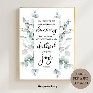 May include: A framed print with a gold border features the text "You turned my mourning into dancing" in elegant script. Eucalyptus leaves in shades of green and gray surround the text. The print includes the words "clothed me with joy" and "Psalm 30:11".