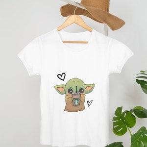 Puede incluir: Camiseta blanca con un personaje de dibujos animados verde que sostiene una taza de caf&eacute;. El personaje tiene ojos grandes y est&aacute; rodeado de corazones.