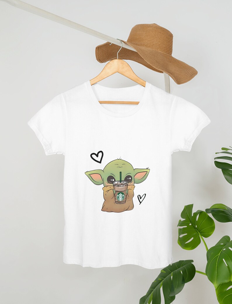 Puede incluir: Camiseta blanca con un personaje de dibujos animados verde que sostiene una taza de caf&eacute;. El personaje tiene ojos grandes y est&aacute; rodeado de corazones.