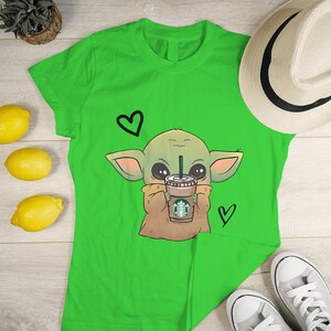 Puede incluir: Camiseta verde con una imagen de dibujos animados de un extraterrestre verde sosteniendo una taza de caf&eacute; con el logotipo de Starbucks. El extraterrestre tiene ojos grandes y est&aacute; rodeado de corazones.