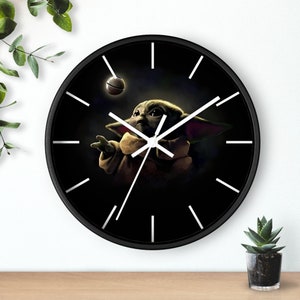 May include: A black wall clock with white hands and numbers. The clock face features a cartoon image of a green alien character from the Star Wars series, The Mandalorian, holding a small silver ball.