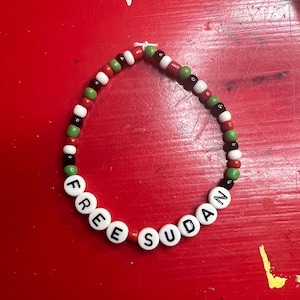 以下が含まれることがあります： 白い文字ビーズで「FREE SUDAN」と書かれたビーズブレスレット。ブレスレットは赤、緑、黒、白のビーズのパターンです。ブレスレットは赤い表面にあります。