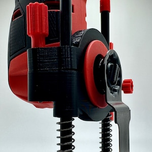 Puede incluir: Un jig de impresión 3D negro y rojo para una herramienta rotatoria con un mecanismo de inmersión de resorte y una cuchilla negra. El jig tiene una base con múltiples agujeros y el texto "NEXTGEN WORKSHOP" está impreso en la base.