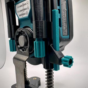 May include: A black 3D printed jig with a spring and a metal blade. The jig is designed to hold a Makita DTM52 18V oscillating tool. The tool is teal and black and has the text "STARLOCK MAX", "STARLOCK PLUS", and "STARLOCK" on the side. The text "Makita DTM52 18V = 10000-20000/min" is also visible on the tool.