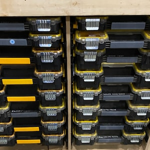 Puede incluir: Un conjunto de contenedores de almacenamiento de plástico negro y amarillo con tapas transparentes. Los contenedores están apilados en dos columnas, con cada columna que contiene 10 contenedores. Los contenedores están etiquetados con diferentes categorías, como "Eléctrico", "Tornillos de hormigón", "Brocas de fresado" y "Brocas de pozo profundo".