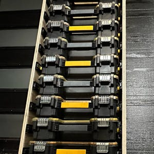 Può includere: Un rack di stoccaggio verticale in legno contenente una pila di dieci organizer per attrezzi in plastica nera. Ogni organizer presenta una maniglia giallo brillante, un coperchio in plastica trasparente e chiusure in metallo.