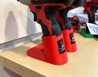 Puede incluir: Dos taladros eléctricos Milwaukee rojos y negros con un soporte de plástico rojo. Los taladros tienen un mango negro y un gatillo plateado. El soporte tiene el logotipo de Milwaukee.