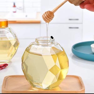 May include: A clear glass honey jar with a faceted design. The jar is filled with honey and has a wooden honey dipper in it. The honey is dripping from the dipper.