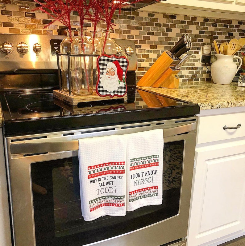 May include: Two white kitchen towels with red and green Christmas-themed designs hang on a silver oven door. The towels have the text "Why is the carpet all wet Todd?" and "I don't know Margo!" printed on them.