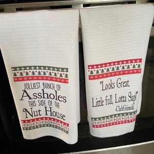 May include: Two white kitchen towels with red and green Christmas-themed borders. The left towel has the text "JOLLIEST BUNCH OF ASSHOLES THIS SIDE OF THE NUT HOUSE". The right towel has the text "Looks Great. Little full, Lotta Sap! -Clark Griswold".