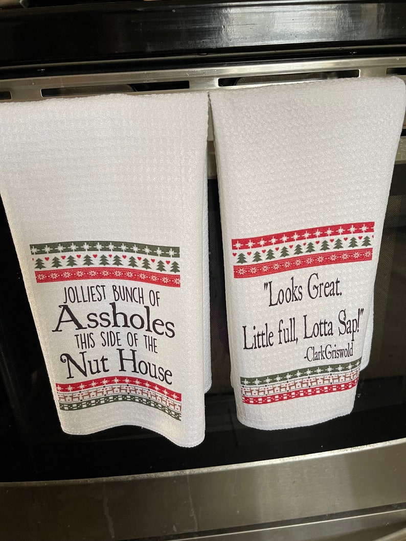 May include: Two white kitchen towels with red and green Christmas-themed borders. The left towel has the text "JOLLIEST BUNCH OF ASSHOLES THIS SIDE OF THE NUT HOUSE". The right towel has the text "Looks Great. Little full, Lotta Sap! -Clark Griswold".