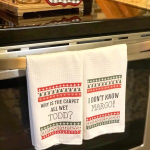 May include: Two white kitchen towels with red and green Christmas-themed borders. The text on the towels reads "Why is the carpet all wet Todd?" and "I don't know Margo!"