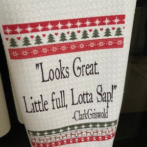 May include: White kitchen towel with a red and green Christmas pattern. The towel has a quote from the movie "Christmas Vacation" that reads "Looks Great. Little full, Lotta Sap!" -Clark Griswold