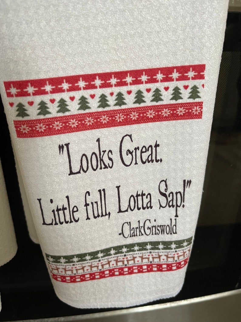 May include: White kitchen towel with a red and green Christmas pattern. The towel has a quote from the movie "Christmas Vacation" that reads "Looks Great. Little full, Lotta Sap!" -Clark Griswold