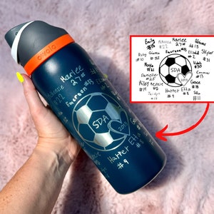 Puede incluir: Una botella de agua azul con tapa blanca y detalles naranjas. La botella está grabada con un balón de fútbol, corazones y el texto "SDA 2013" y "Harper Etta #9".
