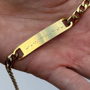 Könnte beinhalten: Ein goldfarbenes Armband mit einem Kettenglied-Design. Das Armband hat eine rechteckige Platte mit Morsecode. Das Armband wird in der Hand einer Person gehalten.