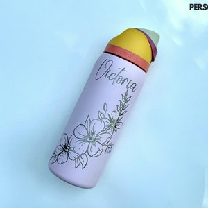 Puede incluir: Una botella de agua reutilizable de color morado claro con un diseño floral y el nombre "Victoria" impreso en ella. La botella tiene una tapa amarilla y naranja.