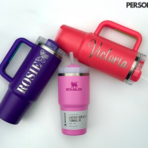 May include: Three stainless steel travel mugs with lids and handles. The mugs are purple, pink, and red. The purple mug has the name "Rosie" engraved on it. The pink mug has the name "Victoria" engraved on it. The red mug has the Stanley logo and the words "Quencher H2O Tumbler" engraved on it.