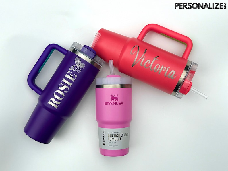 May include: Three stainless steel travel mugs with lids and handles. The mugs are purple, pink, and red. The purple mug has the name "Rosie" engraved on it. The pink mug has the name "Victoria" engraved on it. The red mug has the Stanley logo and the words "Quencher H2O Tumbler" engraved on it.