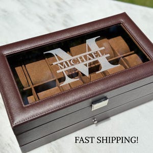 May include: A brown leather watch box with a clear glass top. The box is personalised with the name "Michael" and the letter "M". The interior has brown watch cushions. The box has a silver clasp and a small keyhole.