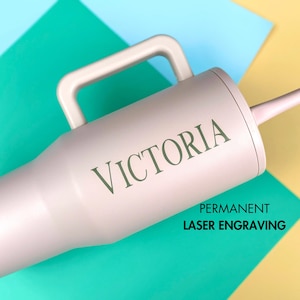 May include: A light pink insulated tumbler with a handle and a straw, featuring the name VICTORIA engraved in dark green lettering. The text PERMANENT LASER ENGRAVING is printed in black below the name on the side of the cylindrical drink container.