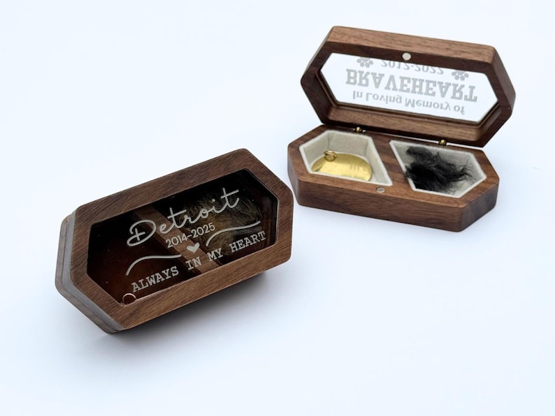 May include: Two wooden keepsake boxes with glass lids. One box is open, revealing a gold pendant and black fur. The other box is closed, with the text "Detroit 2014-2025 ALWAYS IN MY HEART" etched on the glass. The boxes are on a white surface.