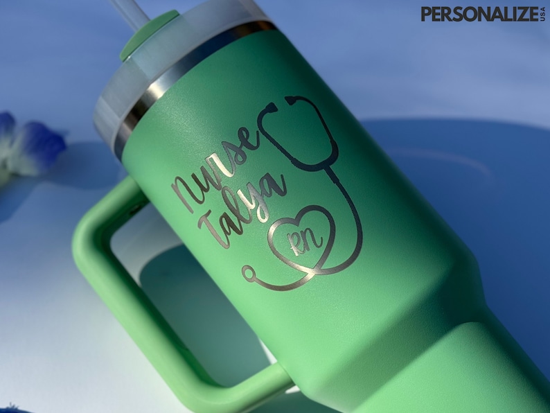 May include: A green travel mug with a handle and a lid. The mug is engraved with the text "Nurse Talya RN" and a stethoscope with a heart.