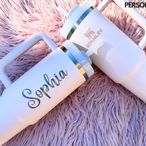 May include: Two white stainless steel travel mugs with handles. One mug has the word "Sophia" written on it in silver lettering. The other mug has the word "Stanley" written on it in pink lettering.