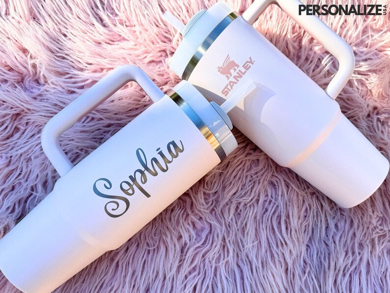 May include: Two white stainless steel travel mugs with handles. One mug has the word "Sophia" written on it in silver lettering. The other mug has the word "Stanley" written on it in pink lettering.