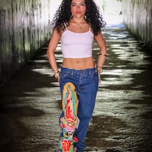 Op de afbeelding: Een persoon draagt een witte crop top en een blauwe spijkerbroek met een kleurrijke religieuze afbeelding op de broekspijp. De jeans wordt gecombineerd met zwarte, rode en witte sneakers. De achtergrond is een donkere tunnel.