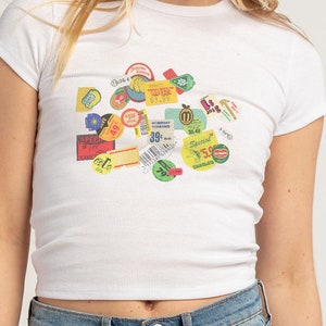 May include: White ribbed crop top featuring a collage of colorful vintage-style price tags and stickers. The tags display various prices and text, including "Special," "Inventory Clearance," and "Banana Square." The top has short sleeves and a crew neck.