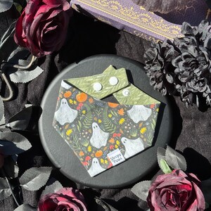 May include: A Halloween-themed pet bandana with a gray background featuring white ghost illustrations, pumpkins, and foliage. The top portion is olive green with white snaps. The bandana is on a black plate, surrounded by dark red roses and a decorative book.